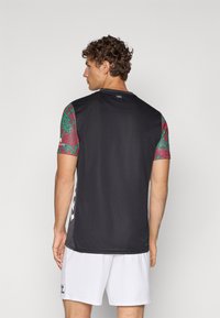 Black athletic shirt with short, colorful patterned sleeves. Features a round neck and "1985" tag at the back. Worn with white shorts.