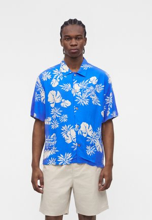 Νέος άνδρας με πλεξούδες στα μαλλιά που φοράει μπλε πουκάμισο με κοντά μανίκια και λευκά τροπικά φύλλα και μπεζ σορτς.