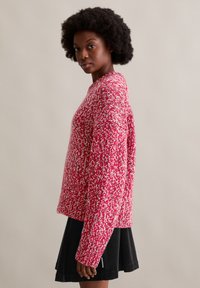 Rot-weißer Strickpullover mit oversized Passform, rundem Ausschnitt und langen Ärmeln, getragen über einem schwarzen, plissierten Rock. Strukturiertes Muster im gesamten Pullover.