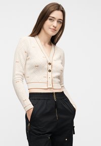 Νεαρή γυναίκα με μακριά καστανά μαλλιά, φορώντας ένα μπεζ cropped ζακέτα με σχέδια και μαύρο ψηλόμεσο παντελόνι με χρυσά φερμουάρ και χέρια στις τσέπες.