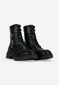 Par de botas de combate de cuero negro con cordones, suelas gruesas y bordado floral gris en los lados exteriores.