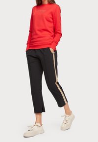 Röd sweatshirt med rund hals och logotyp, tillsammans med svarta byxor med en beige sidostrip och en kortad nederkant. Vita sneakers fulländar looken.