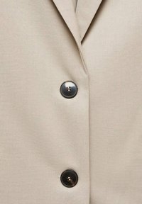 Beiger Stoff mit zwei großen dunklen Knöpfen und einem Kragennaht, was auf eine Nahaufnahme eines geknöpften Kleidungsstücks wie einer Jacke oder einem Mantel hindeutet.