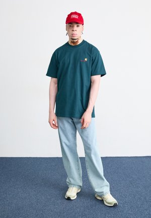 Donkergroen t-shirt met korte mouwen en een logo, lichtblauwe wijde jeans, en beige sneakers. Een rode pet maakt de look compleet.