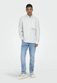 Witte overhemd met dunne strepen, enkele borstzak en tekstdetail aan de zijkant. Lichtblauwe jeans en stevige witte sneakers.