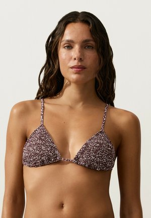 Femeie cu păr lung și ondulat, purtând un top de bikini triunghiular maro și alb cu model, stând în fața unui fundal simplu deschis.