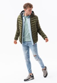 Giubbotto puffo verde oliva con cappuccio, abbinato a una camicia in chambray azzurro chiaro, una t-shirt grigia, jeans blu strappati e sneakers grigie.