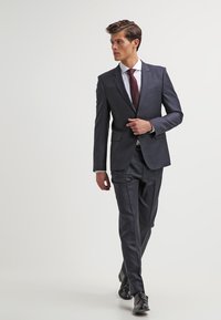 Dark navy suit with a fitted design, matching trousers, a white dress shirt, and a burgundy polka-dot tie. Black dress shoes complete the outfit.