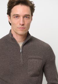 Brauner Reißverschluss-Pullover mit gerippter Textur und einer Brusttasche. Verfügt über einen hohen Kragen und einen kontrastierenden Stoff an der Tasche.