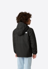 Veste noire imperméable à capuche avec poignets élastiques et logo dans le dos, portée sur un pantalon bleu clair.