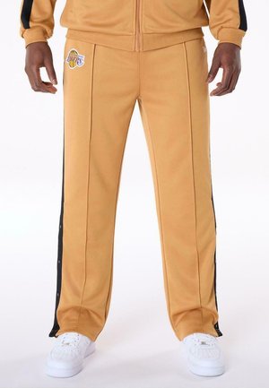 Mann trägt senfgelbe Los Angeles Lakers Trainingshose mit schwarzen Seitenstreifen und weißen Sneakers, steht vor weißem Hintergrund.