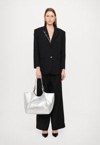 Zwarte oversized blazer met zilveren knoopdetails, gecombineerd met zwarte wijde broek. Houdt een gestructureerde zilveren tote bag vast, pointed zwarte schoenen.