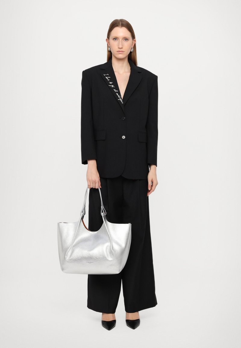 Zwarte oversized blazer met zilveren knoopdetails, gecombineerd met zwarte wijde broek. Houdt een gestructureerde zilveren tote bag vast, pointed zwarte schoenen.