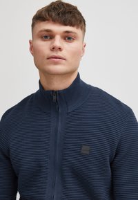 Granatowy sweter na zamek z delikatną prążkowaną fakturą, wysokim kołnierzem oraz małą czarną naszywką z logo po lewej stronie. Miękki materiał i dopasowany krój.