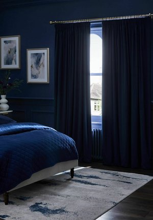 Dunkelblaues Schlafzimmer mit gesteppter Bettdecke, schweren Vorhängen, die am Fenster teilweise geöffnet sind, abstrakter Wandkunst, einer weißen Vase mit Blumen und einem blau gemusterten Teppich.