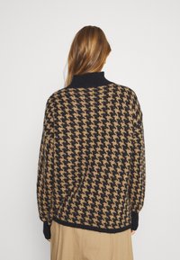 Vrouw met lang bruin haar, gekleed in een zwarte en beige trui met een houndstooth-patroon en beige broek, met haar rug naar de camera tegen een effen achtergrond.