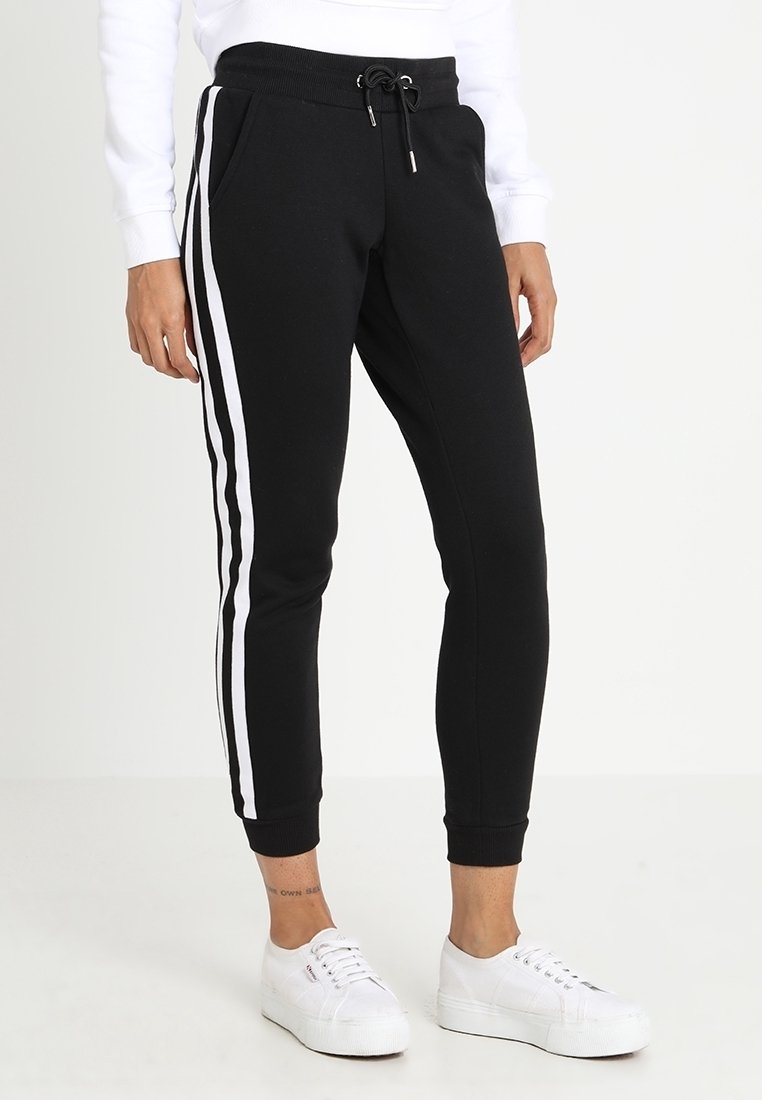 Persoon in zwarte joggers met witte zijstrepen, een wit sweatshirt en witte sneakers, staande op een effen witte vloer en achtergrond.