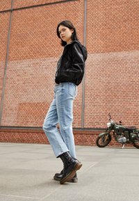 Giacca bomber nera, stampa grafica bianca, jeans blu a gamba dritta, stivali da combattimento neri lucidi con suola spessa, sfondo di muro di mattoni.