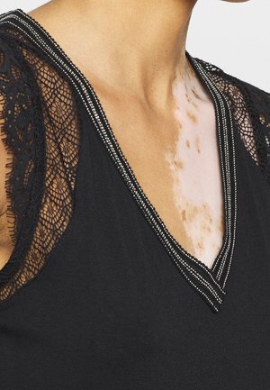 Nahaufnahme von Hals und oberer Brust einer Person mit Vitiligo, die ein schwarzes Oberteil mit Spitzenärmeln und einer mit Perlen besetzten V-Ausschnitt trägt.