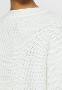 Gros plan sur un pull en tricot blanc côtelé porté par une personne, montrant le haut du dos et les détails de l'encolure.