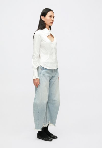 Camisa branca de botões com gola franzida, combinada com jeans de perna larga azul claro com bainha desgastada e sapatos planos pretos.