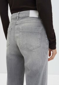 Persoon in hooggeprijsde grijze denim jeans met achterzakken, gecombineerd met een zwart bovenstuk met lange mouwen, tegen een eenvoudige lichte achtergrond.