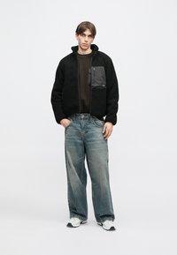 Melna fleeces jaka ar rāvējslēdzēja kabatu krūšu daļā, kombinēta ar vaļīga piegriezuma gaiši zilām džinsa biksēm un baltiem sporta apaviem. Vienkāršs fons.