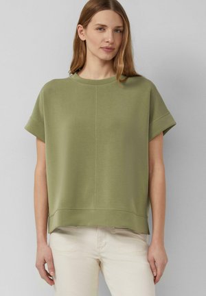 Kobieta o jasnobrązowych włosach, ubrana w zielony, luźny top z krótkim rękawem oraz jasnobeżowe spodnie, stojąca na szarym tle.