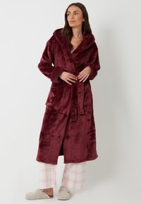 Ropa Marlene en burdeos profundo, hecha de un suave tejido de felpa, con capucha, mangas largas y dos bolsillos frontales. Acompañada de pantalones a cuadros.
