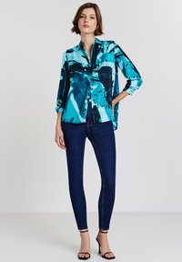 Woman wearing a blue patterned button-up shirt, dark skinny jeans, and black high-heeled sandals standing against a plain background.