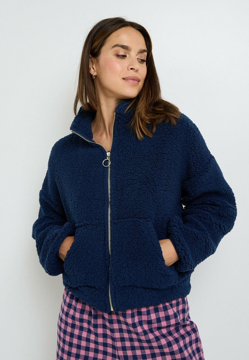 Donna che indossa una giacca sherpa blu navy con zip, con le mani in tasca, abbinata a pantaloni a quadri rosa e blu navy, che guarda di lato.