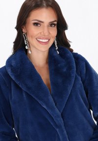 Mørkeblå faux fur frakke med en bred krave, der har en blød tekstur og en omslagedesign. Model iført krystaller øreringe, smilende.