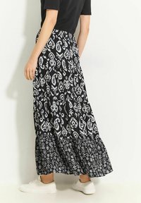 Jupe longue noire à volants, avec un motif cachemire blanc complexe. Confectionnée en tissu léger, portée avec des baskets blanches.