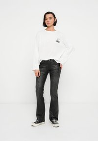 Λευκό μακρυμάνικο πουκάμισο με γραφικό λογότυπο, συνδυασμένο με μαύρα τζιν με φαρδιά μπατζάκια και μαύρα αθλητικά παπούτσια. Απλός σχεδιασμός, casual εφαρμογή, ορατή υφή υφάσματος.