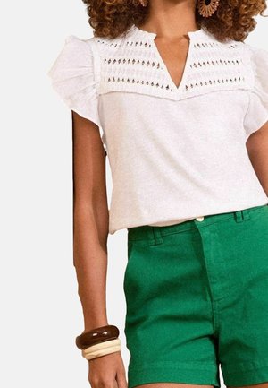 Vrouw draagt een witte blouse met kanten details en pofmouwen, groene high-waisted shorts, en dikke bruine en beige armbanden.