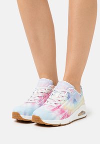 Nogi w pastelowych, tie-dye sneakersach Skechers z białymi sznurowadłami, wyściełaną podeszwą i brązowymi gumowymi elemętami na białym tle.