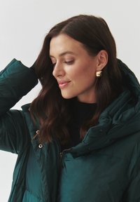 Tyrkysová péřová bunda s vysokým límcem, stříbrnými cvoky a zipem. Modelka má na sobě zlaté náušnice a černý top, s vlnitými hnědými vlasy.