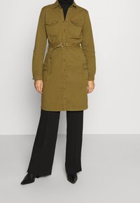 Abito camicia verde oliva con maniche lunghe, chiusura a bottoni frontale, tasche al petto, cintura in vita e tessuto testurizzato; abbinato a pantaloni neri e tacchi.