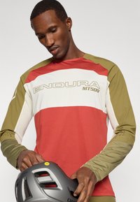 Maglia da ciclismo a maniche lunghe di colore rosso, verde oliva e crema con pannelli in rete. Logo "ENDURA MT500" in evidenza. Tiene un casco grigio.
