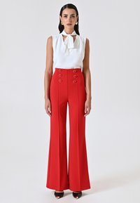Débardeur blanc sans manches avec un détail de col noué, associé à un pantalon évasé rouge taille haute avec des boutons ronds doubles et une texture lisse.