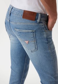 Jeans de denim azul claro con un diseño ajustado, que presentan un parche de cuero marrón con el logotipo de GUESS y dos bolsillos traseros con costura en forma de triángulo.