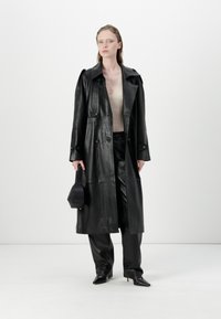 Black leather trench coat with buttons, side pockets, and a wide collar, paired with black pants and a matching handbag. Textured knit top visible.