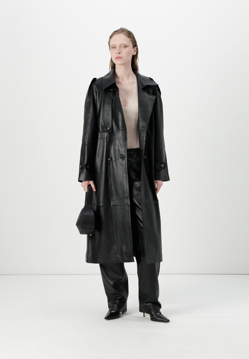 Schwarzer Leder-Trenchcoat mit Knöpfen, Seitentaschen und breitem Kragen, kombiniert mit schwarzen Hosen und einer passenden Handtasche. Strukturiertes Strickoberteil sichtbar.