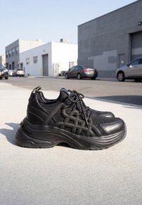 Paire de chaussures de sport noires avec des détails en mesh et cuir, posées sur un trottoir urbain ensoleillé avec des voitures garées et des bâtiments industriels à proximité.