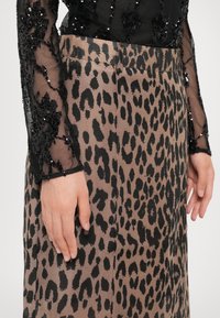 Mains reposant sur les côtés, portant des manches longues transparentes à sequins noirs et une jupe marron avec un motif léopard noir.