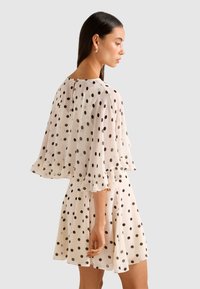 Woman in a white dress with black polka dots, featuring flared sleeves and a ruffled hem, standing in profile against a gray background.