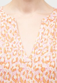 Λαιμός γυναίκας με ανοιχτό ροζ μπλούζα σε σχήμα V και αφηρημένο πορτοκαλί και λευκό λουλουδάτο μοτίβο.