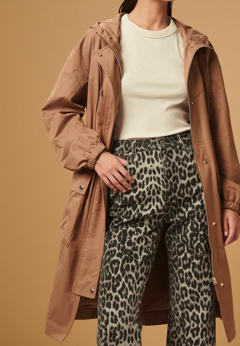 Woman wearing light beige long-sleeve top, brown hooded coat, and high-waist leopard print pants against a beige background.