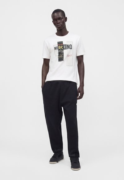 Άνδρας που φορά λευκό T-shirt με σχέδιο, μαύρο παντελόνι και μαύρα αθλητικά παπούτσια, στέκεται με το ένα χέρι στην τσέπη, μπροστά σε απλό φωτεινό φόντο.