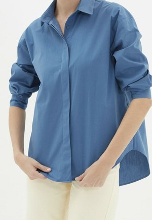 Persoon draagt een los blauw overhemd met knopen en opgerolde mouwen en een lichte broek, terwijl hij de voorkant van het overhemd aanpast.